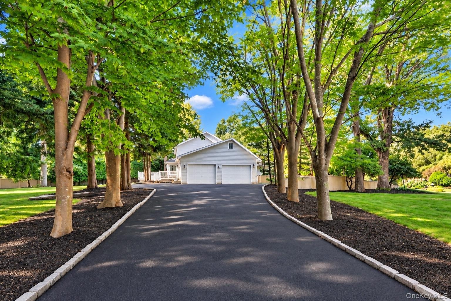Asphalt driveway bordered by stone pavers, flanked by mature trees and manicured lawns, leading to a two-car garage