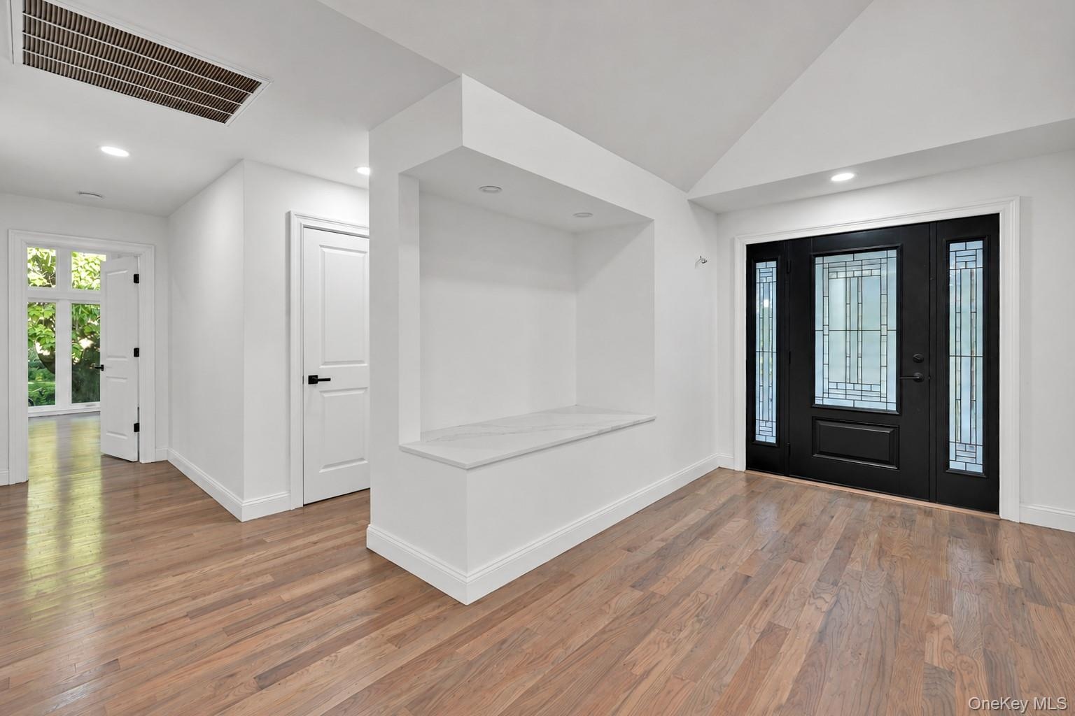 Entryway featuring wood-finish flooring, a built-in recessed display niche with a solid surface bench, vaulted ceiling, and a black-framed front door 