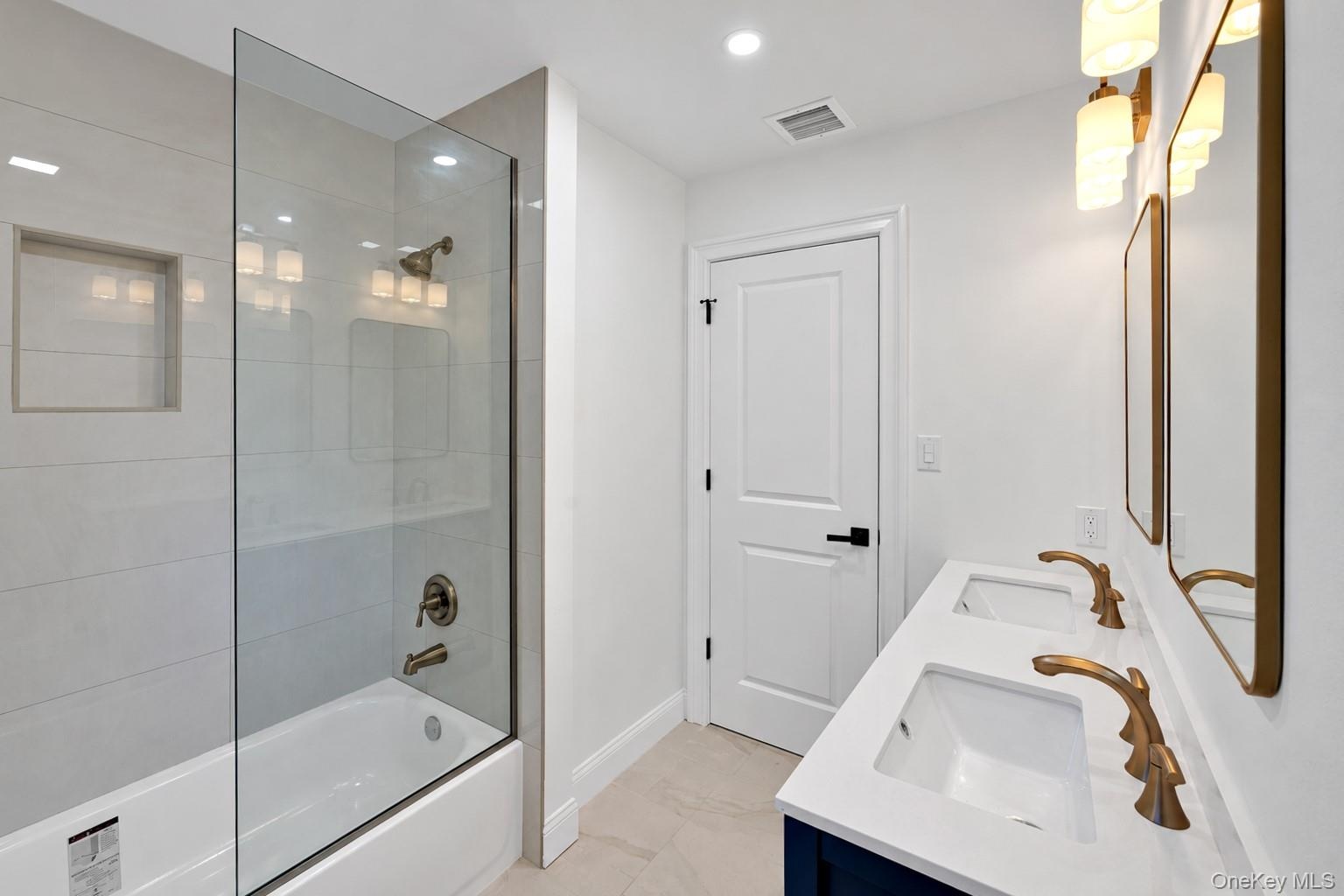Double vanity with rectangular undermount sinks, stone-finish countertops, brass-toned fixtures, and a glass-enclosed tub-shower combination with inte
