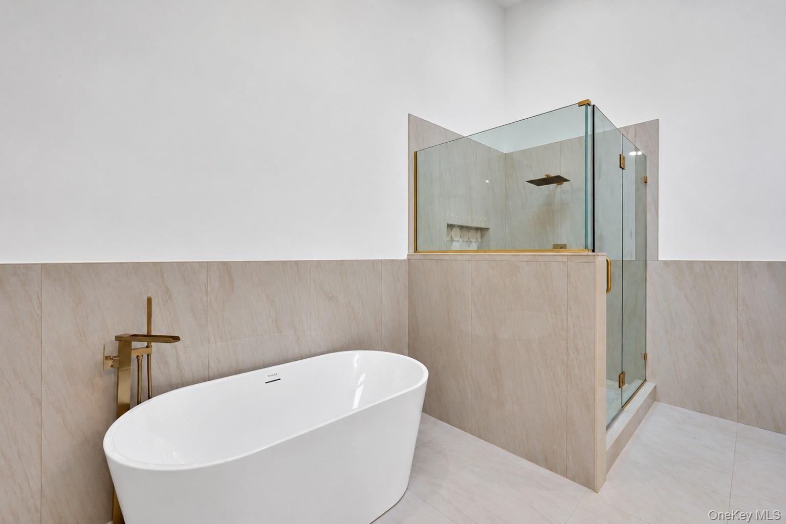 Freestanding white soaking tub with gold-tone floor-mounted faucet, walk-in glass shower enclosure, gold-tone shower fixtures, and neutral large-forma