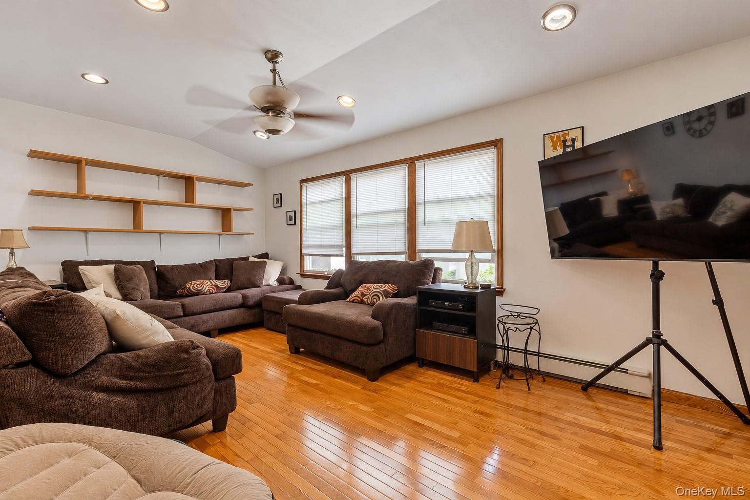 Living area featuring wood-finish flooring, recessed lighting, a ceiling fan, tiered built-in shelving, and multiple windows with wood-trimmed casings