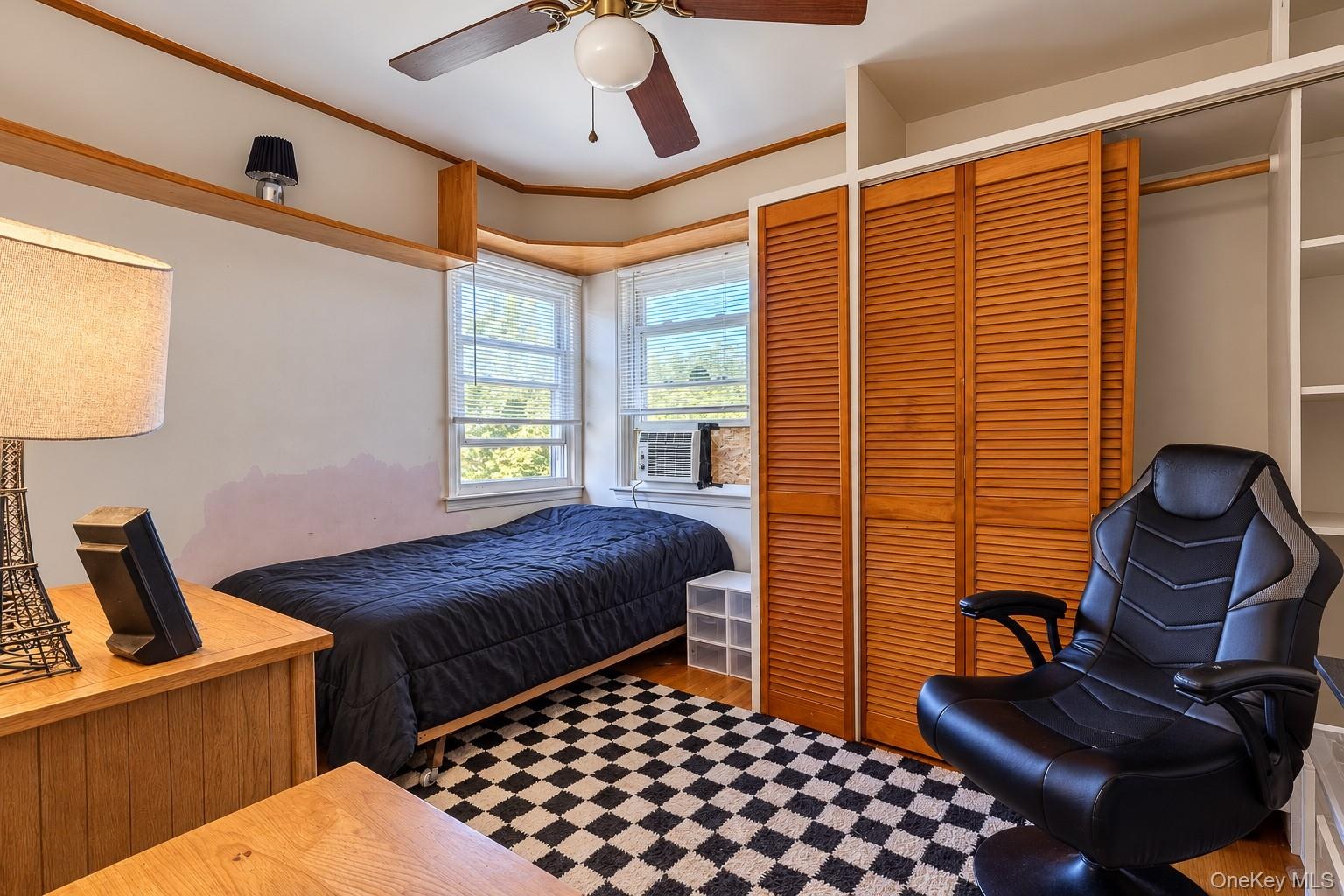 Bay window, built-in wooden shelving, dual-panel louvered closet doors, and a ceiling fan with integrated lighting