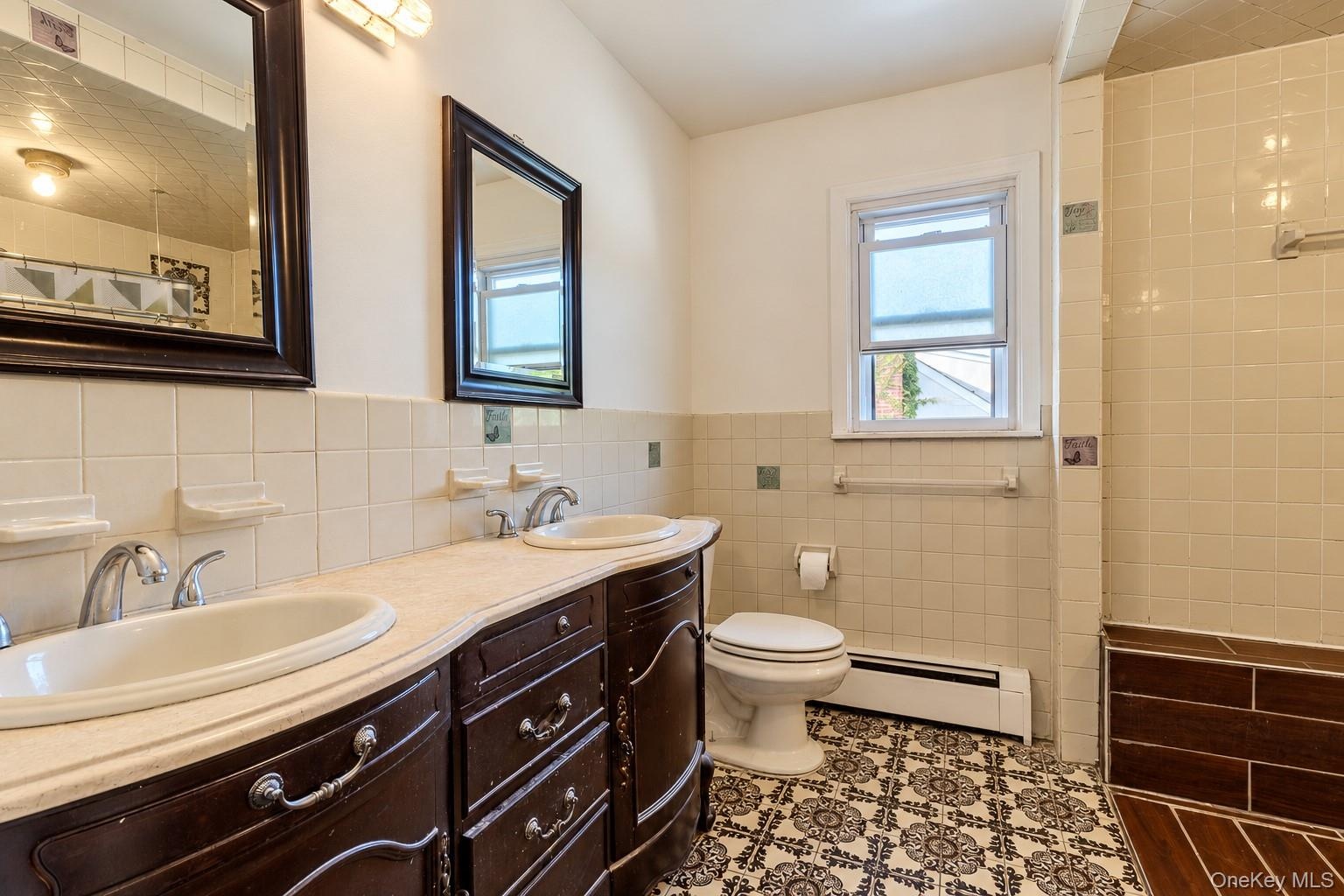 Bathroom featuring a dual-sink vanity with light-toned countertop, ornate dark-finish cabinetry, decorative tile flooring, and a tiled shower surround