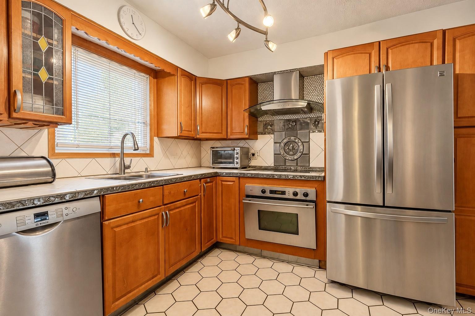 Kitchen featuring custom wood cabinetry, stainless steel appliances, a modern chimney-style range hood, hexagonal tile flooring, and a decorative tile