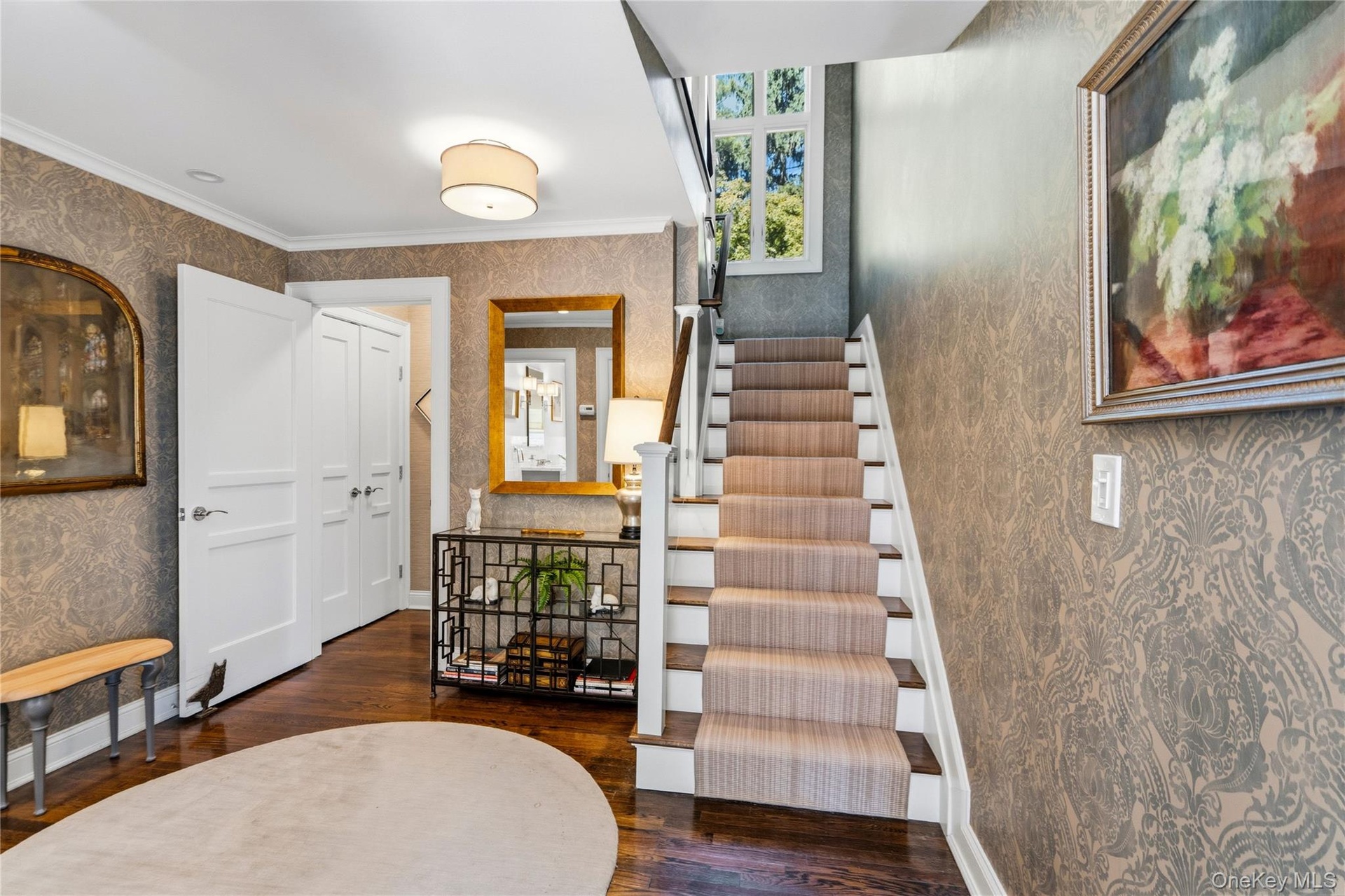 Foyer featuring patterned wallpaper, dark-stained wood flooring, a staircase with a central runner, a multi-pane fixed window, and crisp white millwor