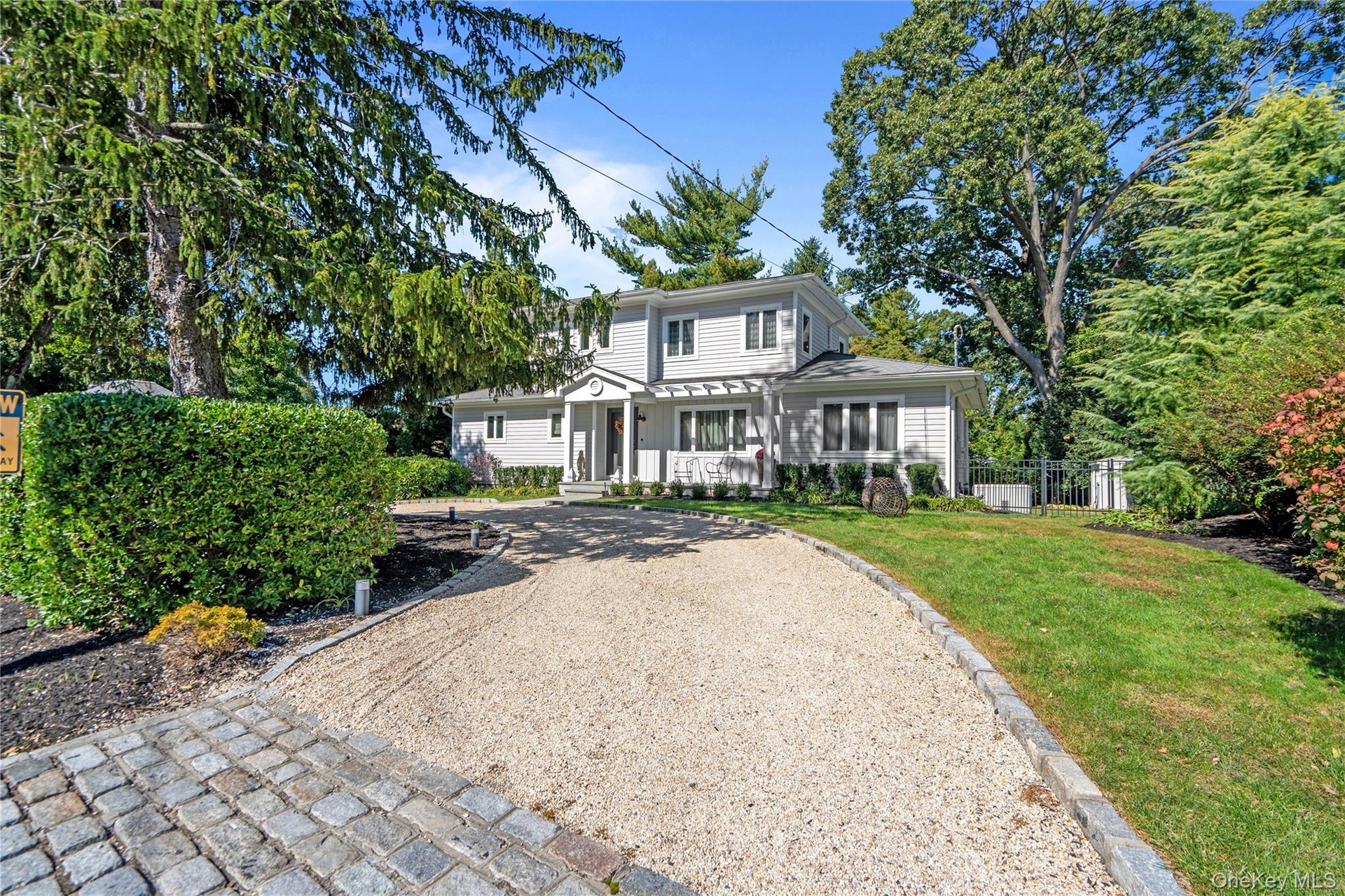 Two-story exterior featuring light-toned horizontal siding, a covered entryway with a pediment, a gravel driveway, cobble-style apron, and mature land
