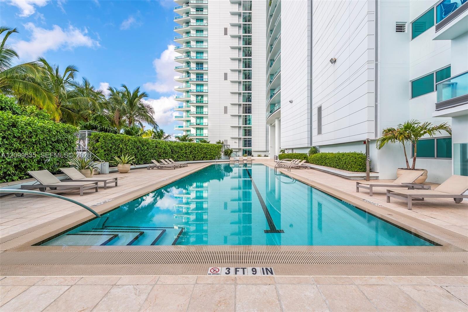 Lap pool with integrated steps, surrounded by a stone-tiled deck and lush green landscaping, set against a modern high-rise building