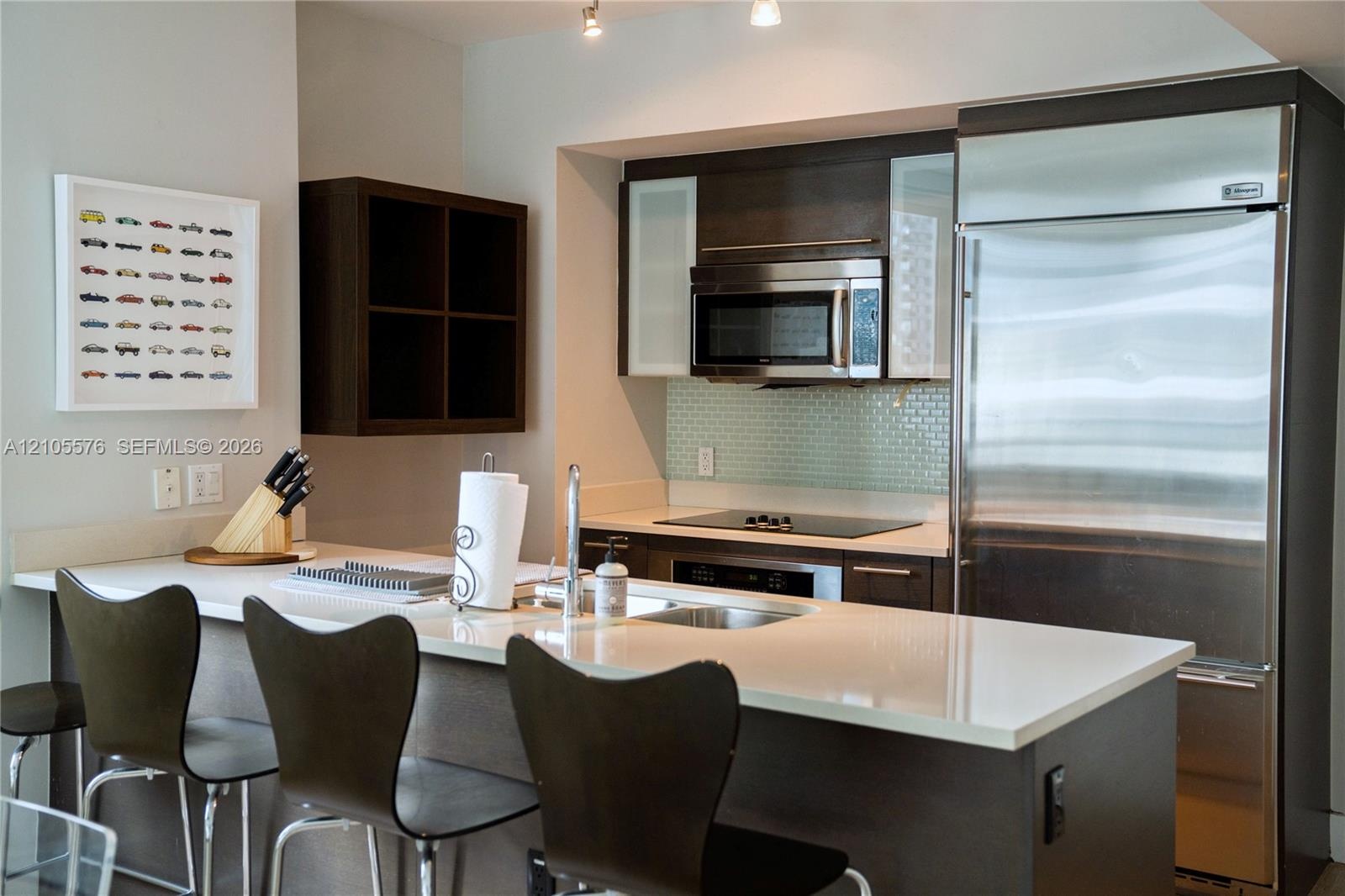 Kitchen featuring dark wood-finish cabinetry, light-toned stone countertops, a glass tile backsplash, stainless steel appliances, and an integrated br