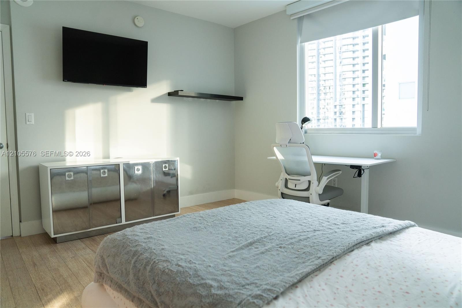 Light-filled room featuring wood-finish flooring, a large window with roller shade, and a wall-mounted floating shelf