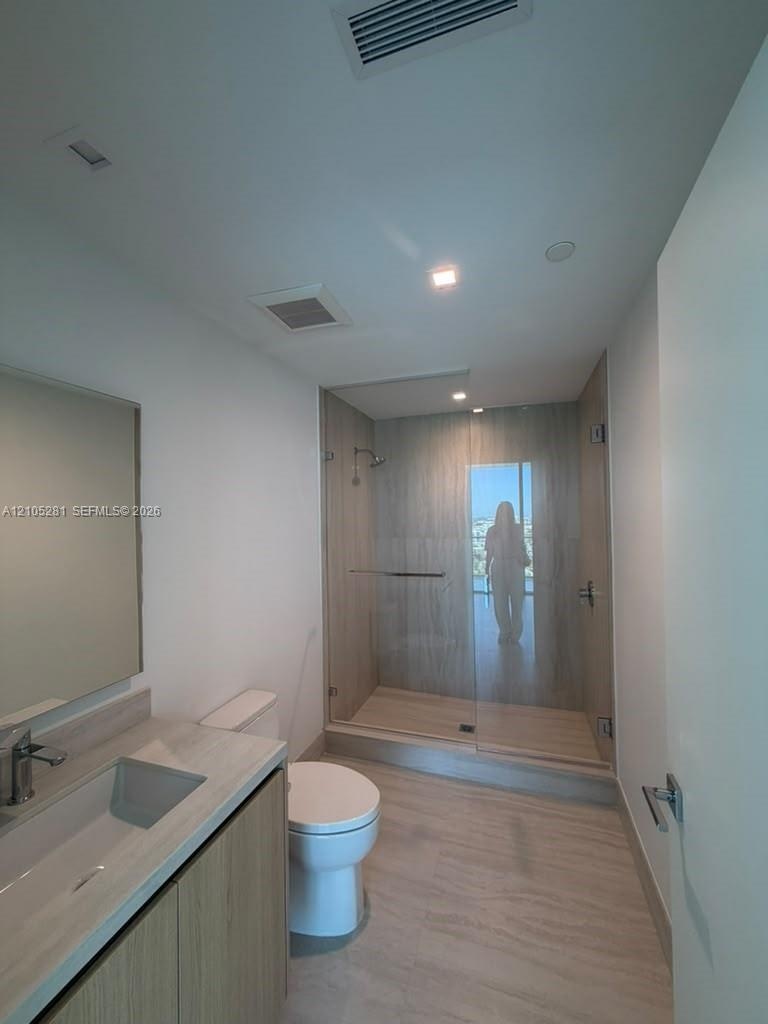 Bathroom featuring a wood-finish vanity with a neutral stone countertop, frameless glass shower enclosure with floor-to-ceiling tile, and integrated r