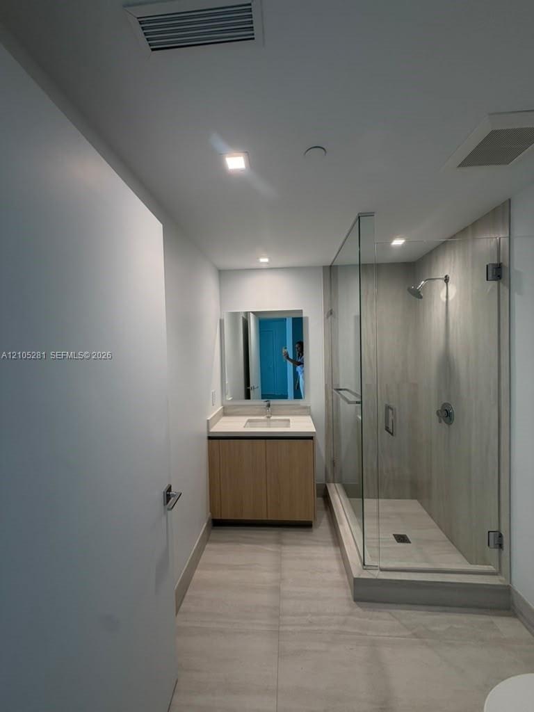 Bathroom featuring wood-finish cabinetry, a rectangular undermount sink, a glass-enclosed shower with full-height tile surround, and large-format floo
