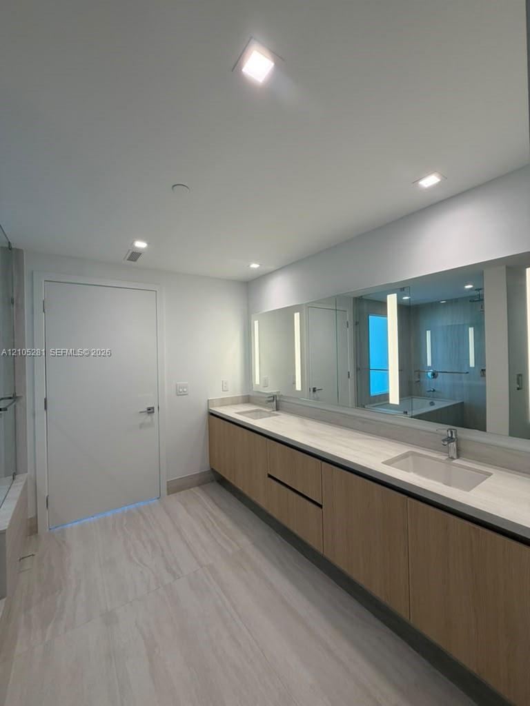 Double vanity featuring light-toned wood cabinetry, dual undermount sinks, stone-finish countertops, wall-to-wall integrated mirror with vertical LED 