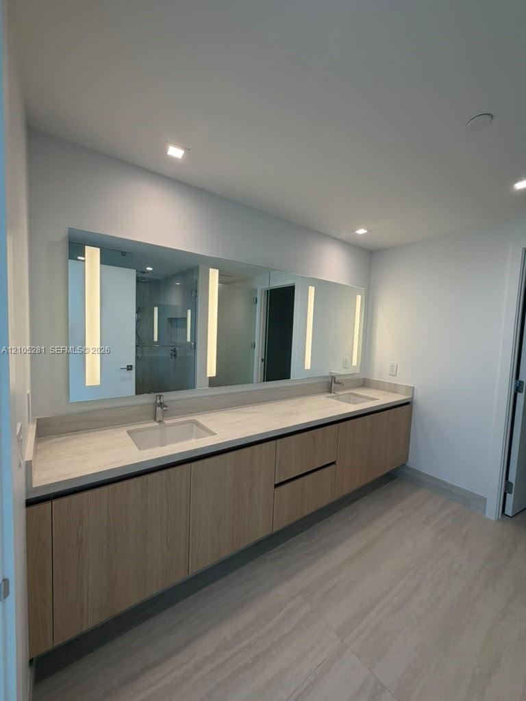 Floating vanity featuring light wood-finish cabinetry, a full-width mirror with integrated LED lighting, and a dual-sink stone-composite countertop
