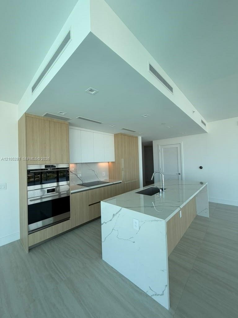 Modern kitchen featuring a waterfall-edge island, composite stone countertops, flat-panel wood-finish cabinetry, stainless steel wall-mounted oven and