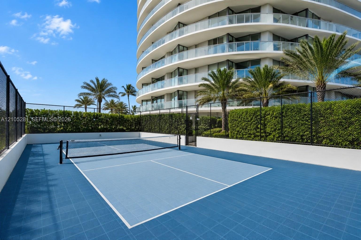 Outdoor pickleball court featuring a multi-toned blue modular surface, perimeter chain-link fencing, and white masonry boundary walls