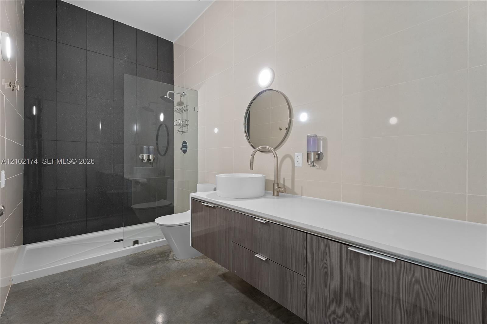 Floating vanity with a white vessel sink, polished countertop, circular mirror, and floor-to-ceiling dark tile accent wall