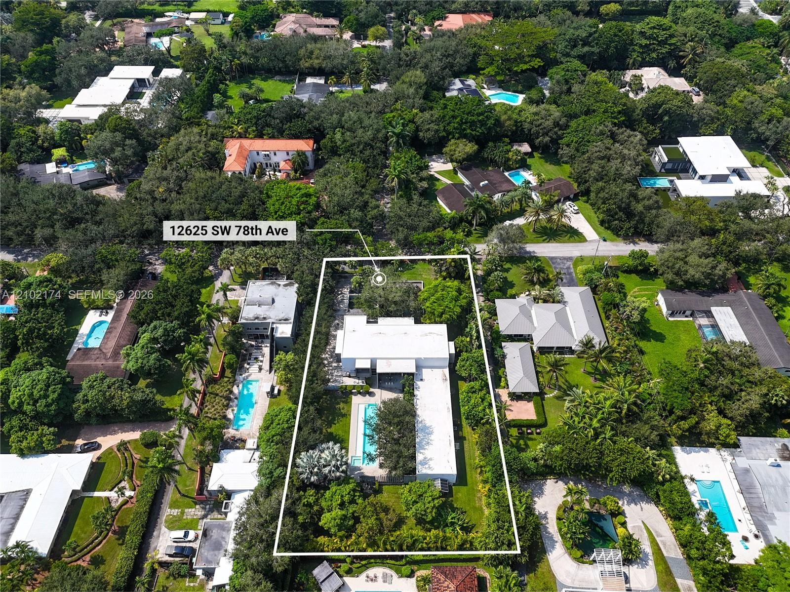 Aerial perspective of a property featuring a rectangular swimming pool, expansive manicured lawn, mature tree canopy, and multi-level roof structure