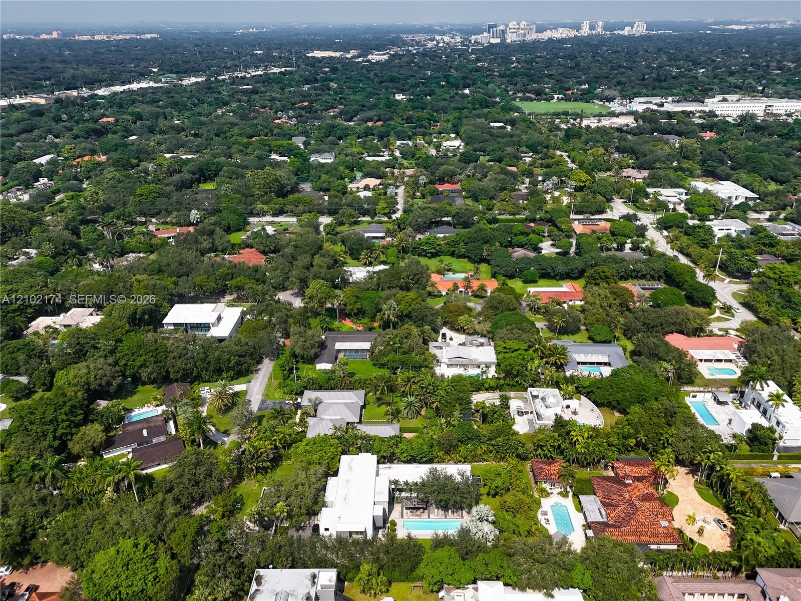 Aerial perspective featuring a flat-roofed estate with a rectangular swimming pool, expansive green lawn, mature trees, and paved driveway