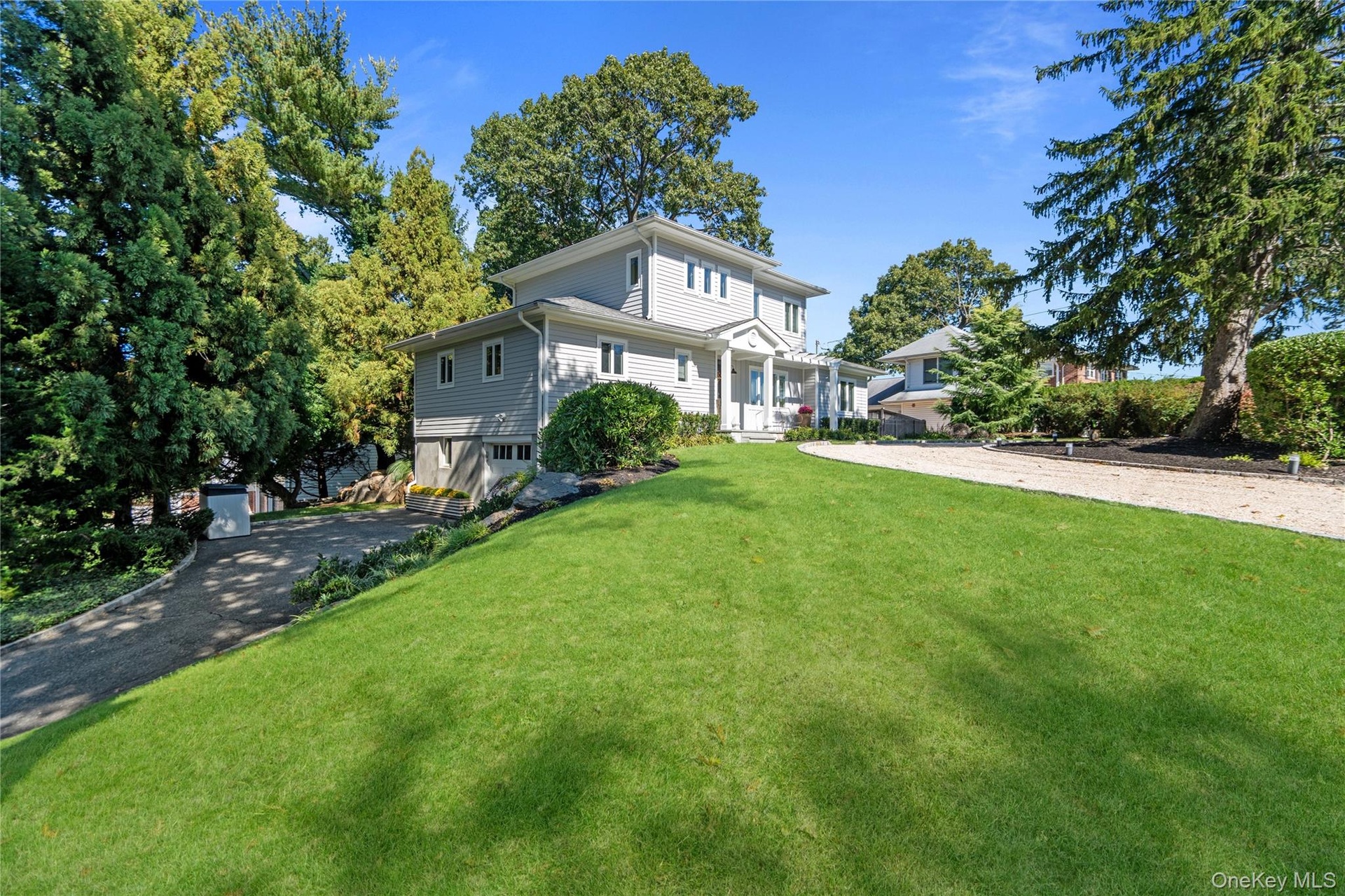 Two-story residence featuring grey horizontal lap siding, a front entry portico with dual columns, a gravel driveway, and mature landscaping
