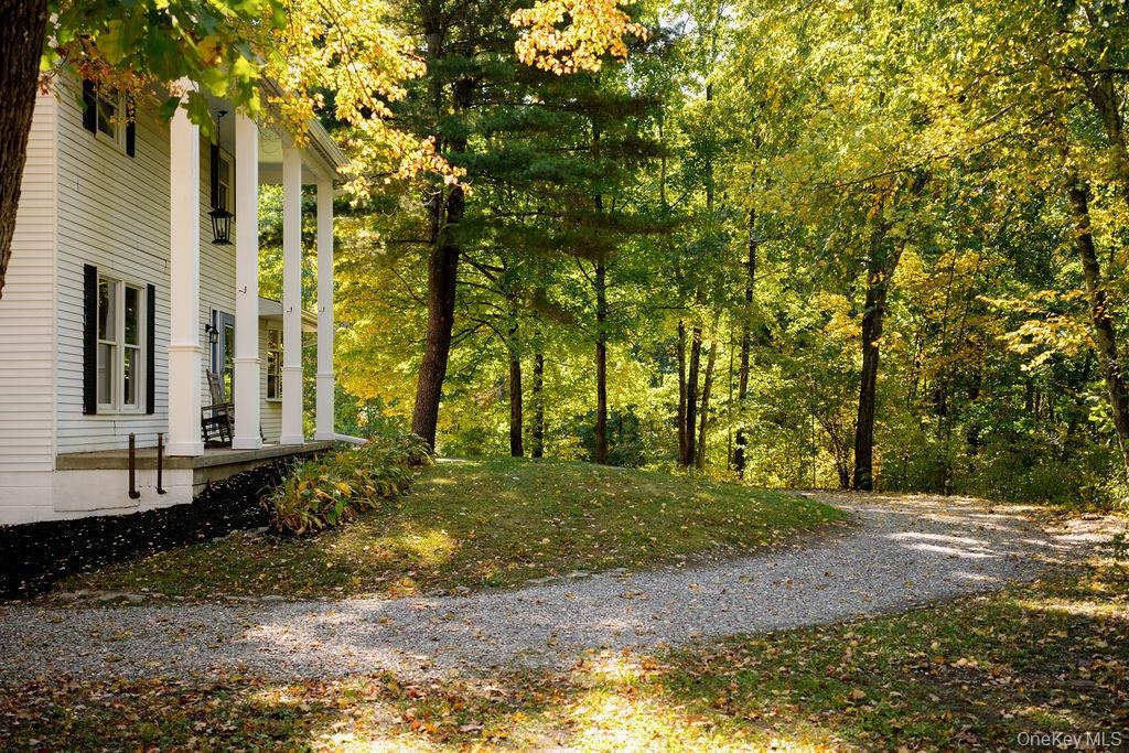 Two-story clapboard exterior featuring a covered front porch, structural columns, shuttered windows, and a gravel driveway set amidst mature trees