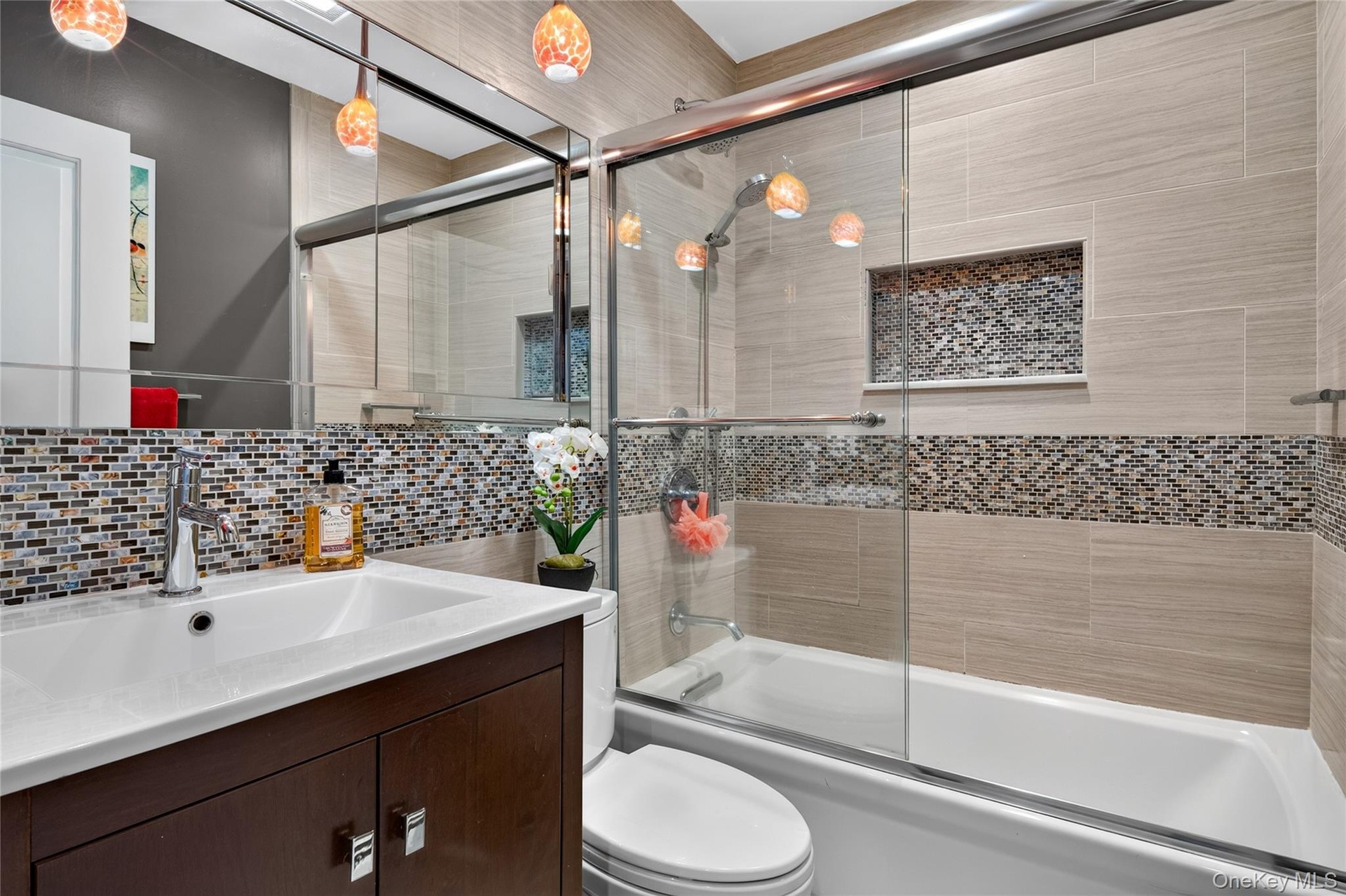 Bathroom featuring a vanity with an integrated rectangular sink, glass-enclosed shower and tub combination, multi-colored mosaic tile accents, wall-mo