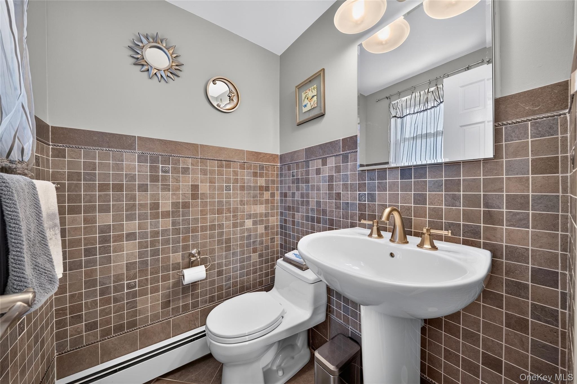 Pedestal sink with a brass-finish widespread faucet, square tile wainscoting, large frameless wall mirror, and dual-bulb vanity light fixture