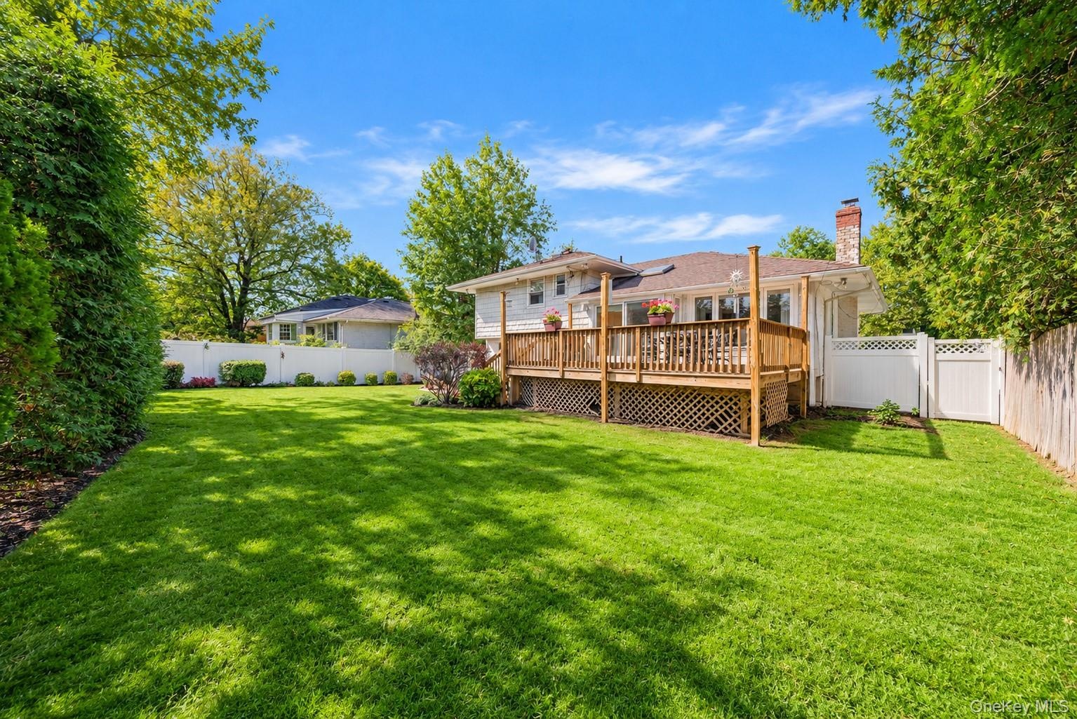 Expansive backyard featuring a manicured lawn, raised wooden deck with lattice skirting, brick chimney, and privacy fencing