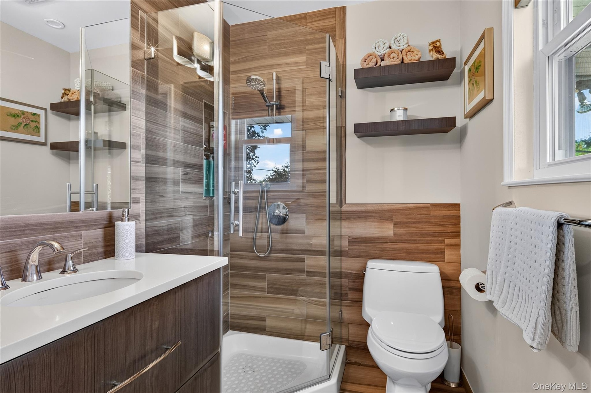 Bathroom featuring a walk-in shower with glass enclosure, wood-finish wall tile, white solid-surface countertop with integrated basin, dark wood-finis
