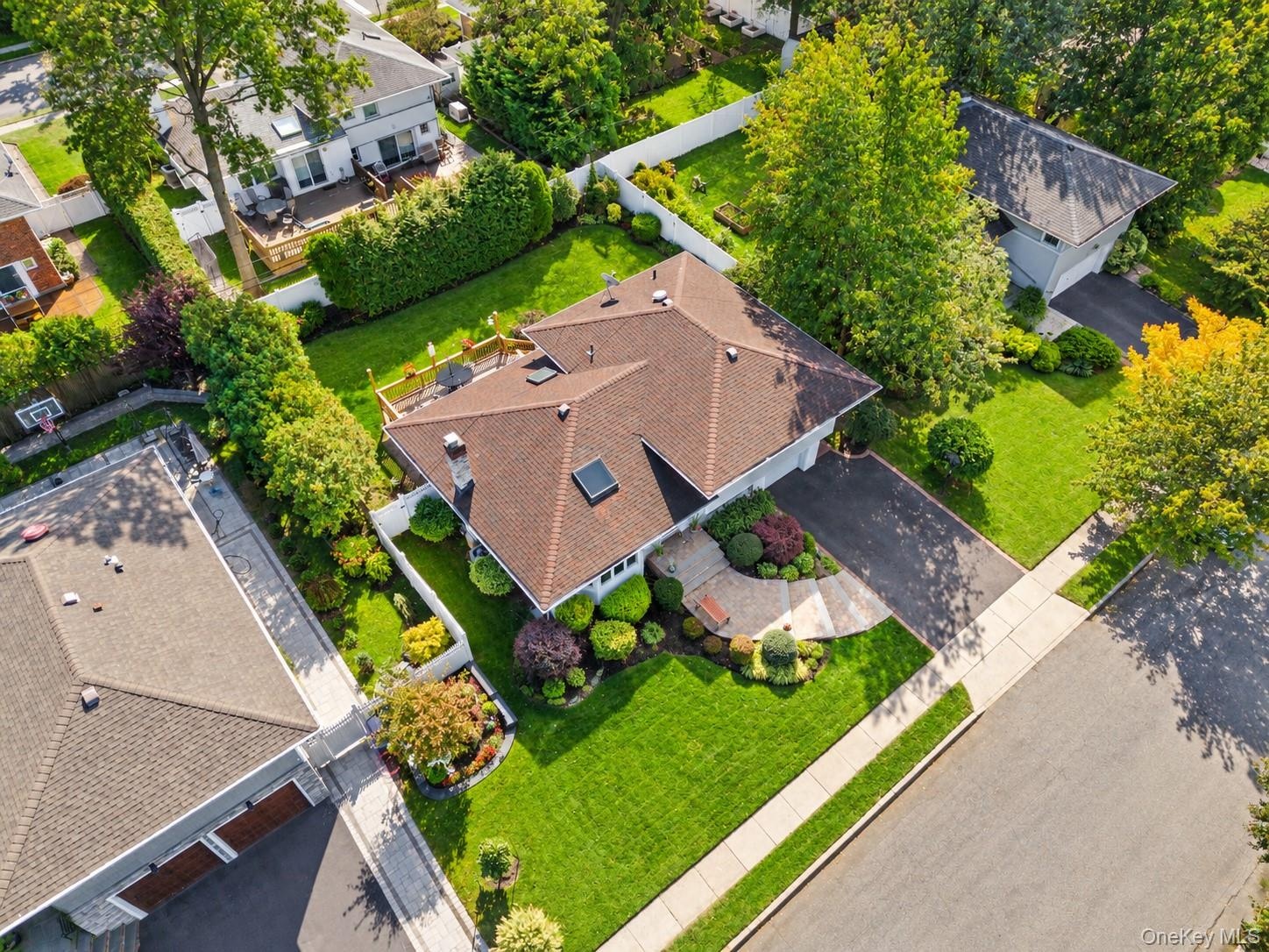 Aerial perspective of a residence featuring a brown shingle roof with a chimney and skylights, an elevated rear deck, a paved driveway, and mature lan