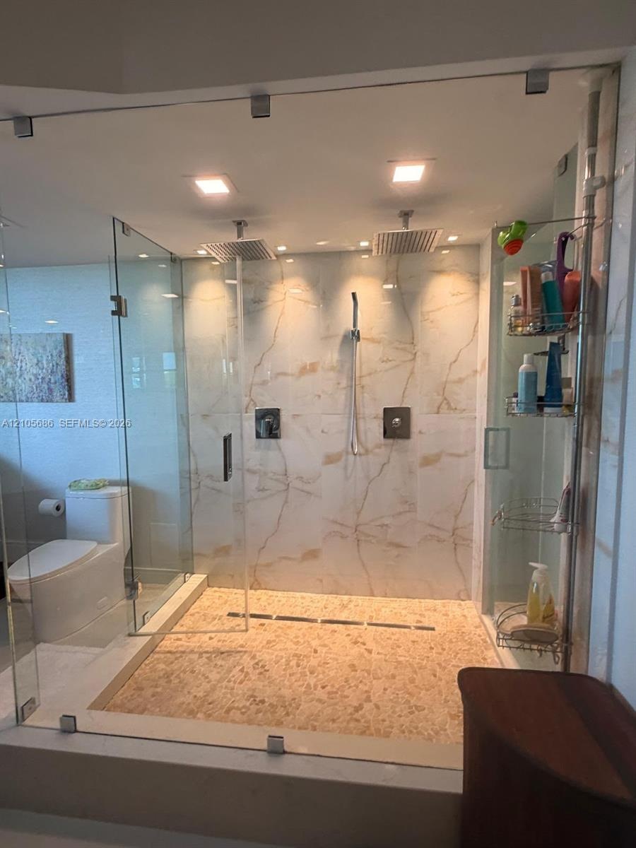 Walk-in shower featuring a frameless glass enclosure, dual rain-style showerheads, hand-held sprayer, large-format stone-look wall tile, and a texture