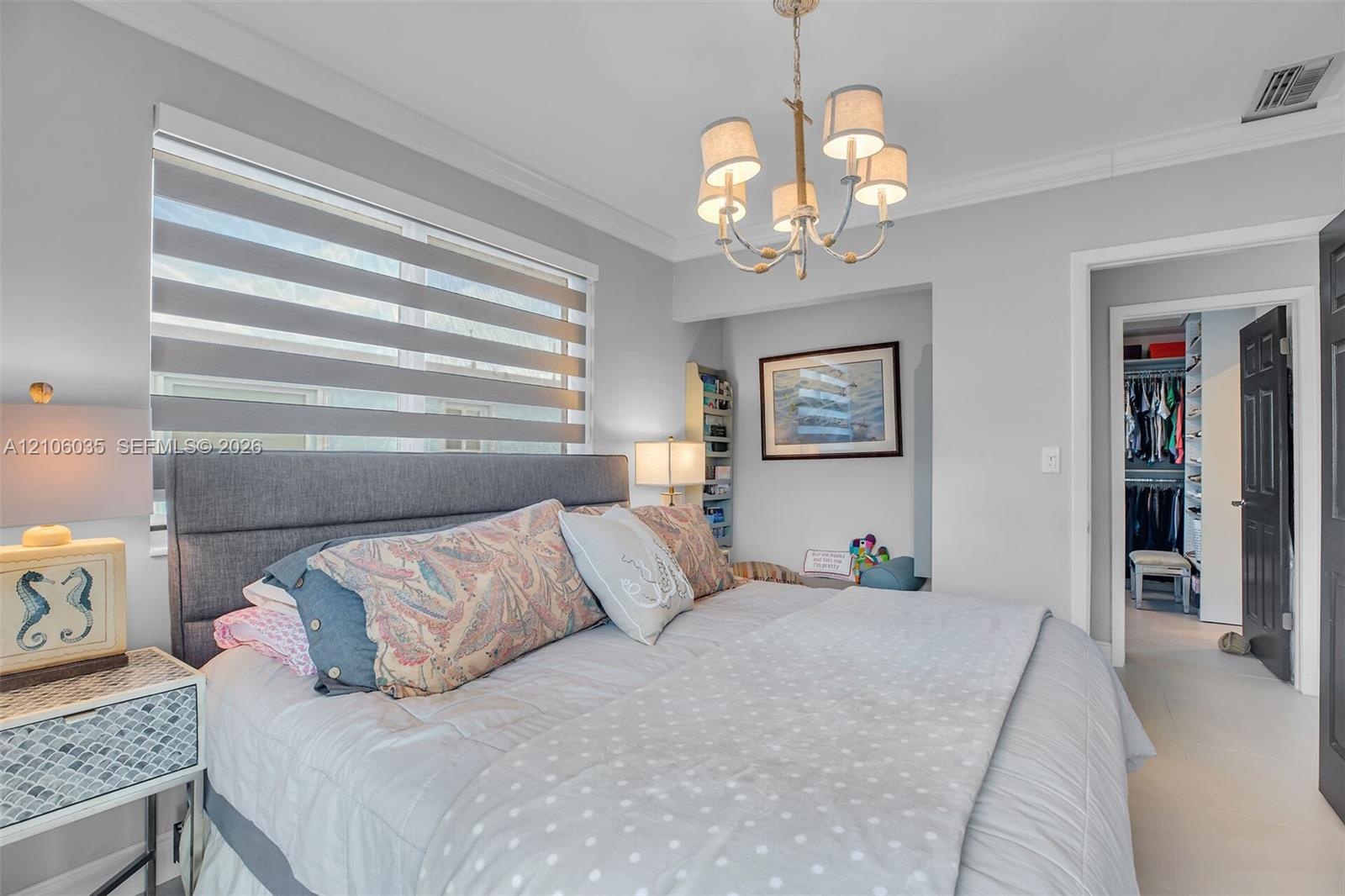 Neutral-toned bedroom featuring crown molding, a five-arm metal chandelier, and layered dual-fabric window shades