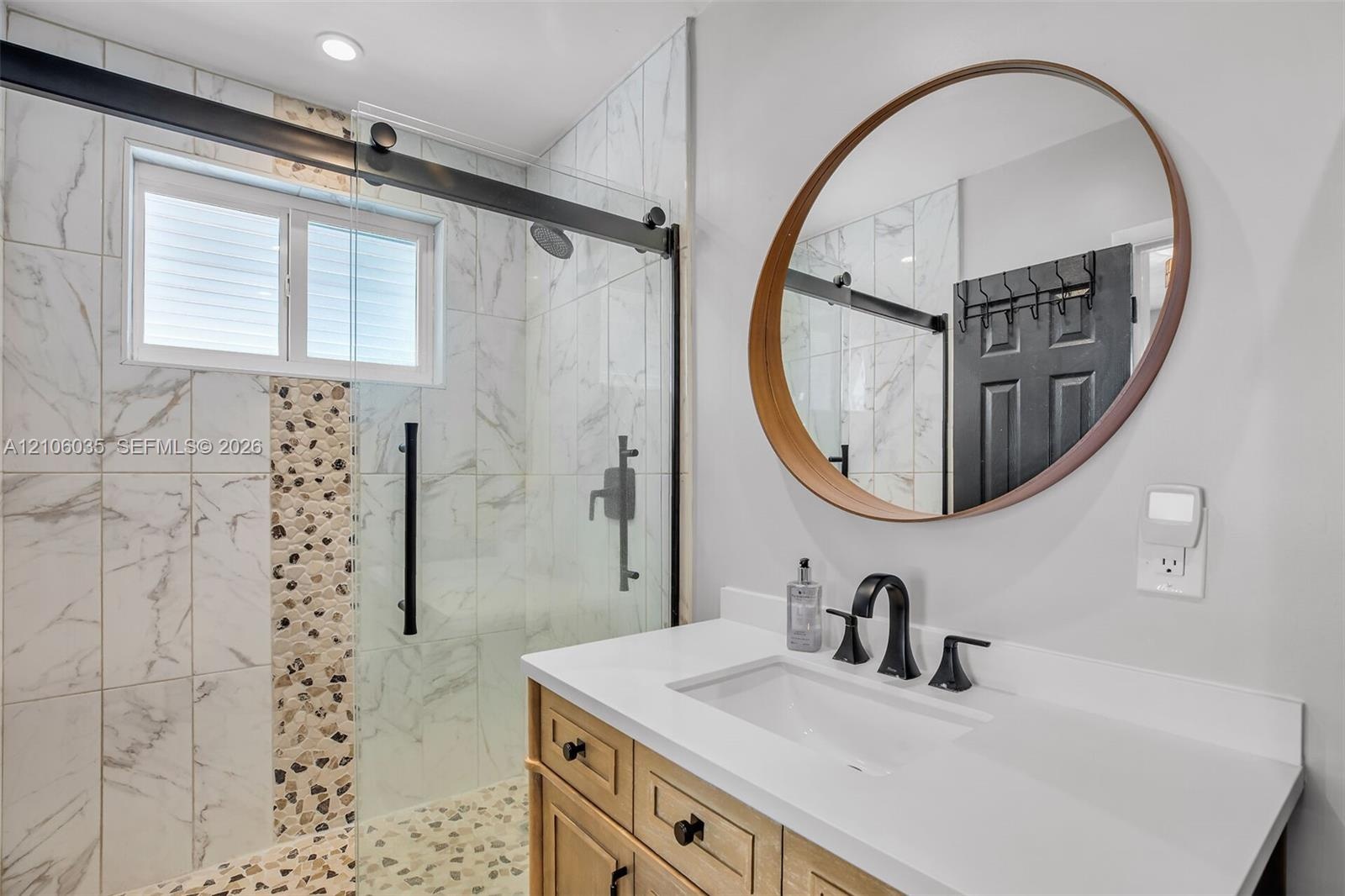 Bathroom featuring a wooden vanity with a white countertop, circular wood-framed mirror, and a walk-in shower with a sliding glass door, patterned sto
