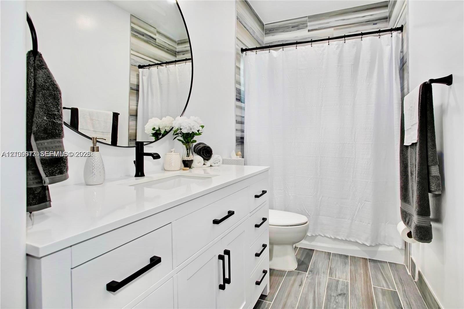 White vanity with solid-surface countertop and matte black hardware, integrated sink, circular mirror, and wood-finish tile flooring