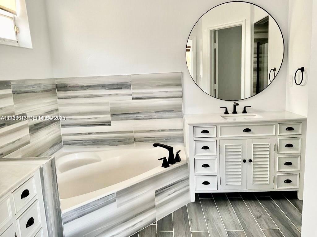 White vanity with louvered cabinet doors, quartz countertop, corner soaking tub with matte black faucet, and horizontal wall tile surround