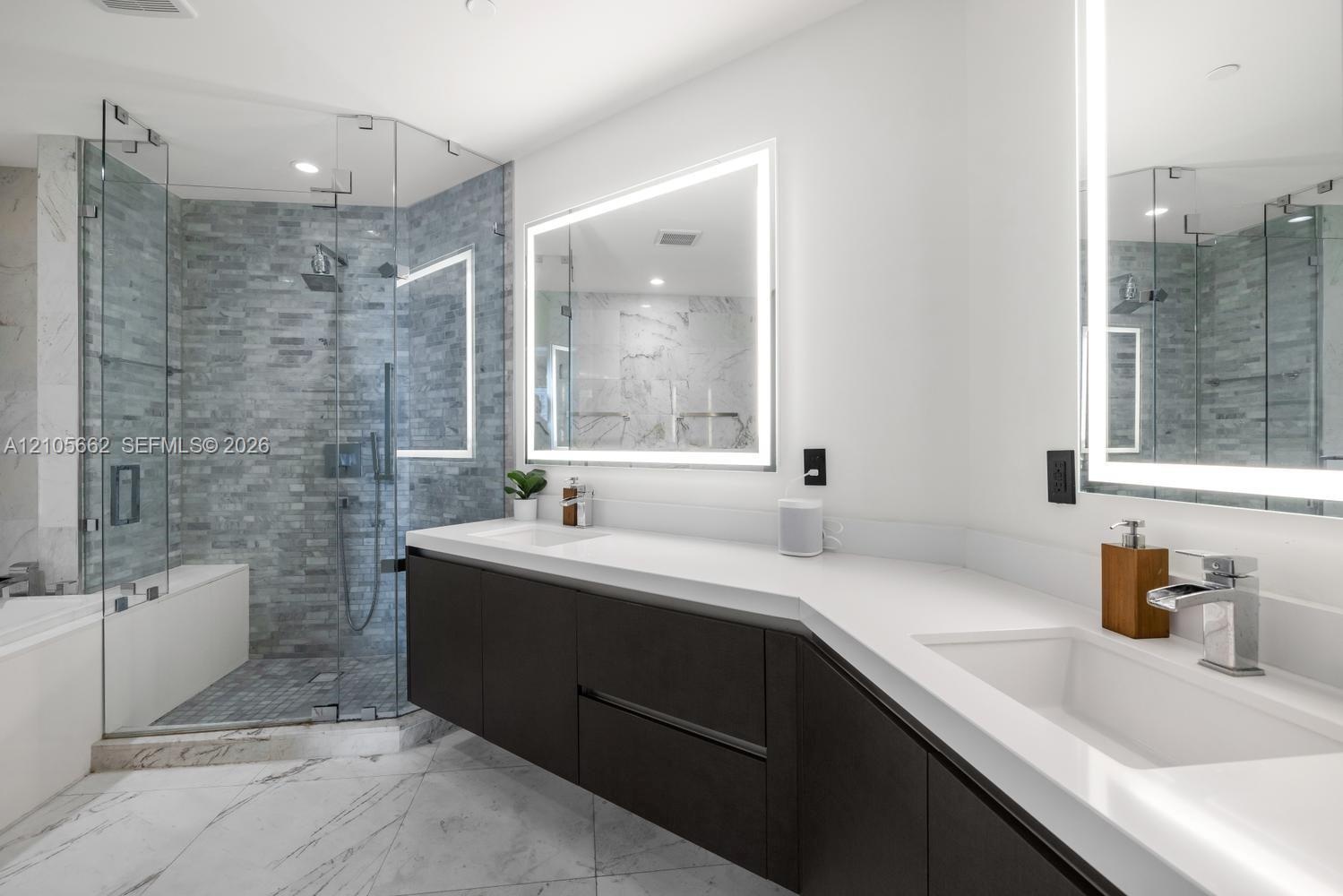 Floating dual-sink vanity with matte dark cabinetry and white countertops, glass-enclosed walk-in shower with textured tile surround, and dual LED-bac