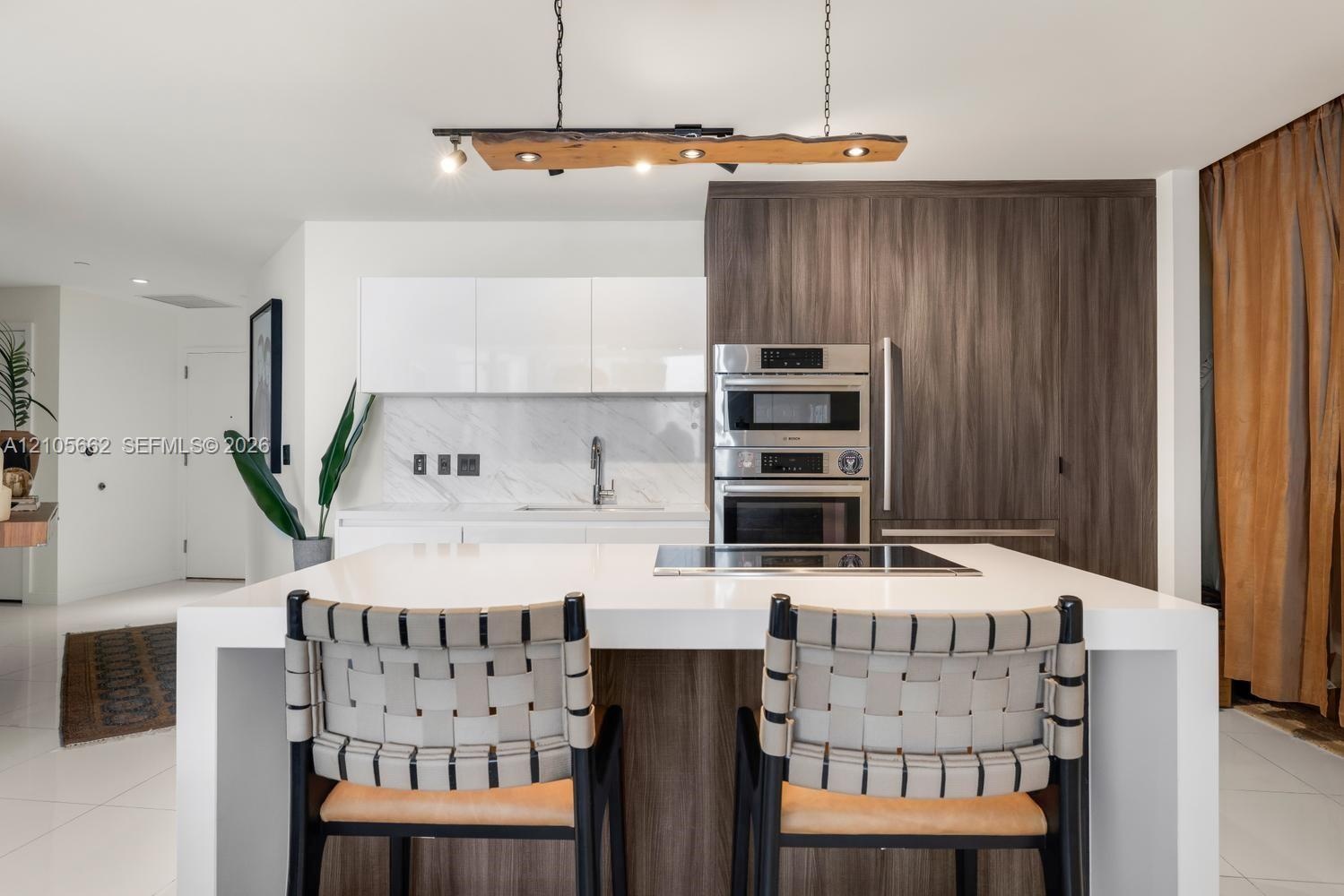 Kitchen featuring a waterfall-edge center island, built-in stainless steel double oven, custom wood-finish cabinetry, natural stone-patterned backspla