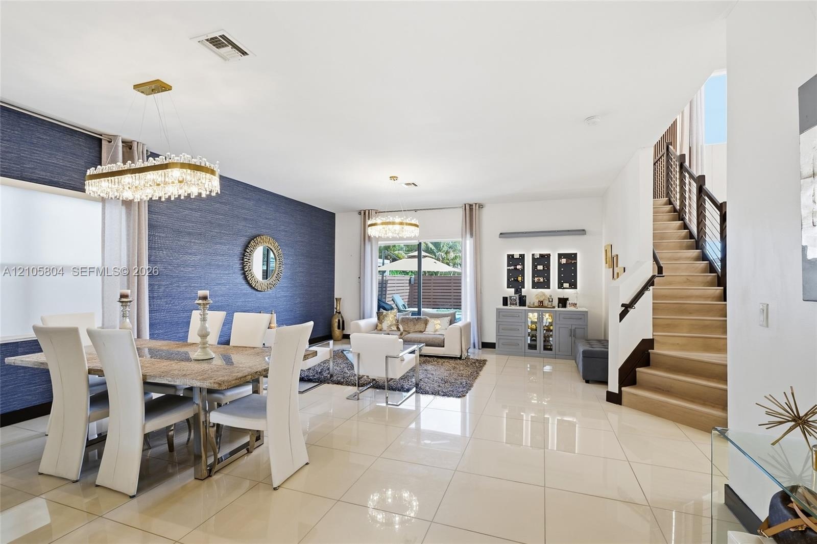 Open-concept interior featuring oversized tile flooring, a blue textured accent wall, dual crystal chandeliers, and a staircase with wood-tread stairs