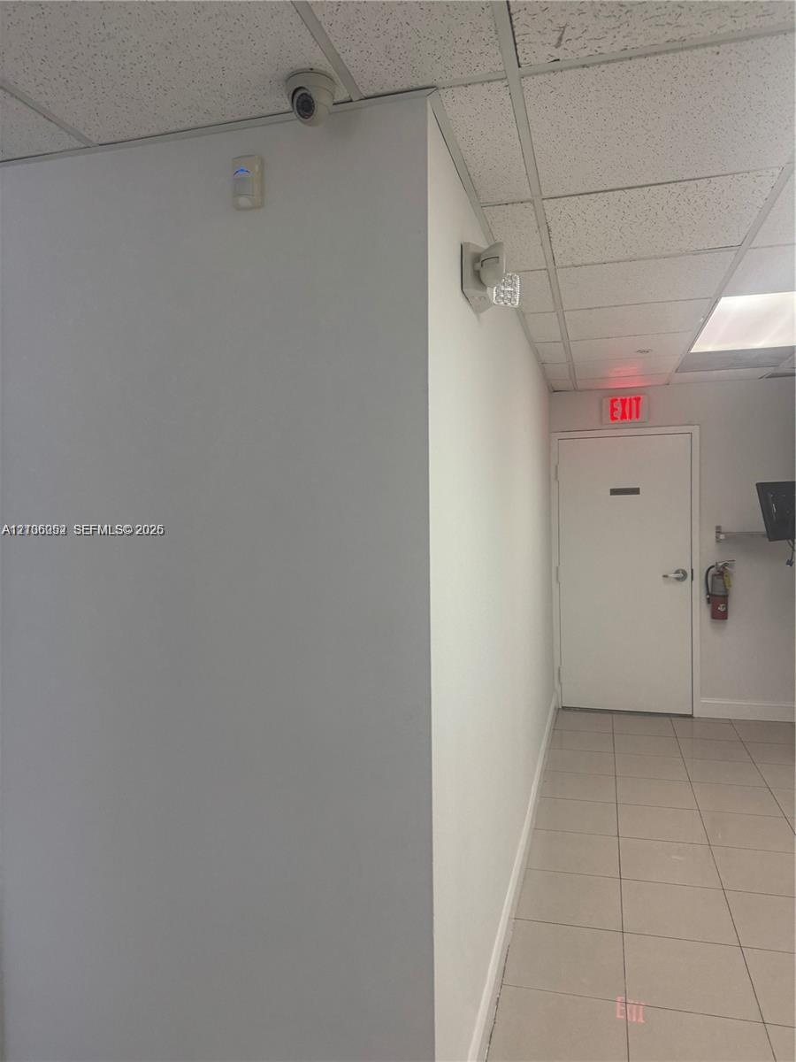 Interior space featuring neutral-toned tile flooring, drop ceiling with recessed lighting, integrated security camera, and wall-mounted exit signage