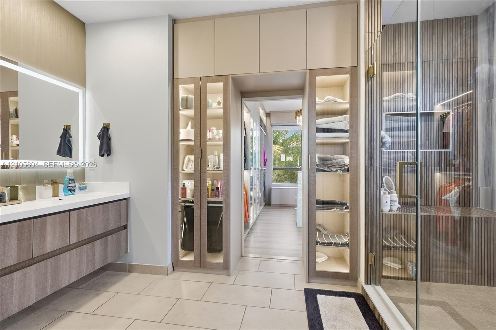 Floating vanity with stone countertop and backlit mirror, floor-to-ceiling built-in cabinetry featuring glass-front display panels, and a walk-in show