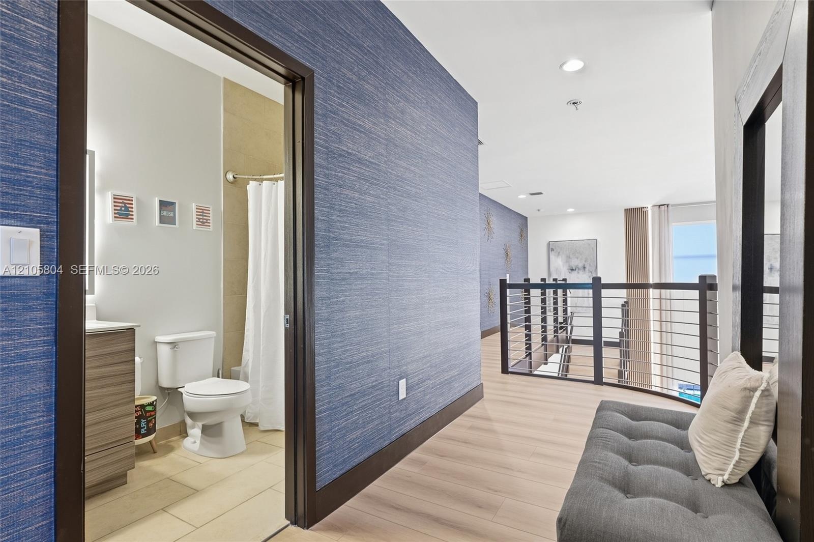 Textured blue wall covering, wood-finish flooring, metal railing with horizontal bars, and bathroom featuring a vanity with integrated sink