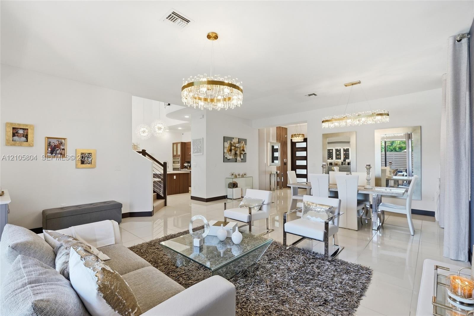 Open-concept living space featuring high-gloss tile flooring, dual tiered crystal chandelier fixtures, and a glass-paneled staircase railing