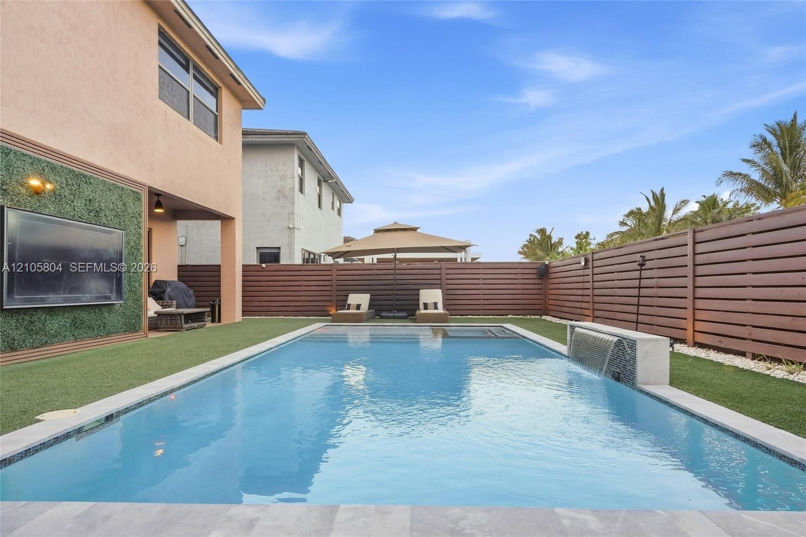 Backyard featuring a rectangular swimming pool with a built-in water feature, horizontal wood-slat privacy fencing, artificial turf lawn, and a wall-m