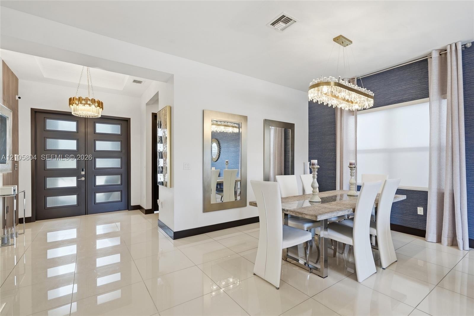 Dining area featuring large-format tile flooring, textured wall covering, custom linear crystal chandelier, and double front entry doors with frosted 