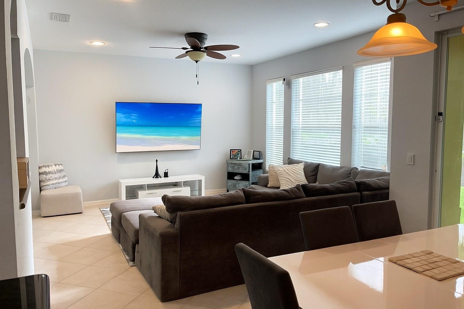 Living area featuring neutral tile flooring, recessed ceiling lighting, a multi-blade ceiling fan, and three large windows with horizontal blinds