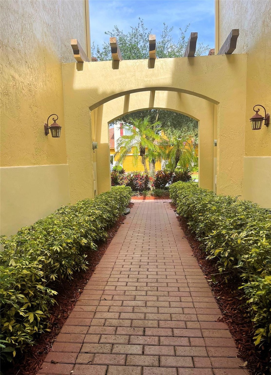 Brick paver walkway lined with manicured hedges, featuring a stucco archway, decorative wood-beam accents, and wall-mounted exterior sconces
