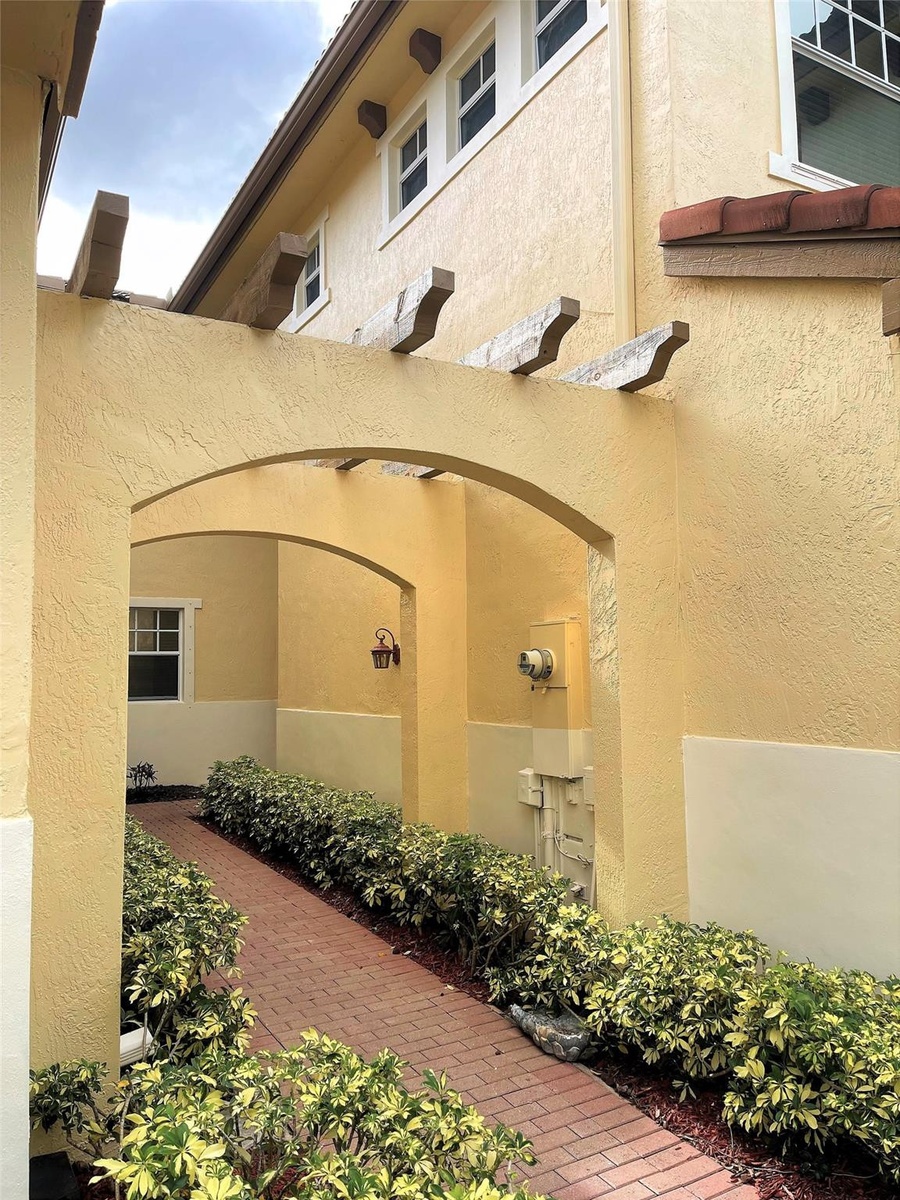 Stucco exterior featuring dual arched walkways, exposed timber rafters, and a brick-paved path bordered by mature shrubbery