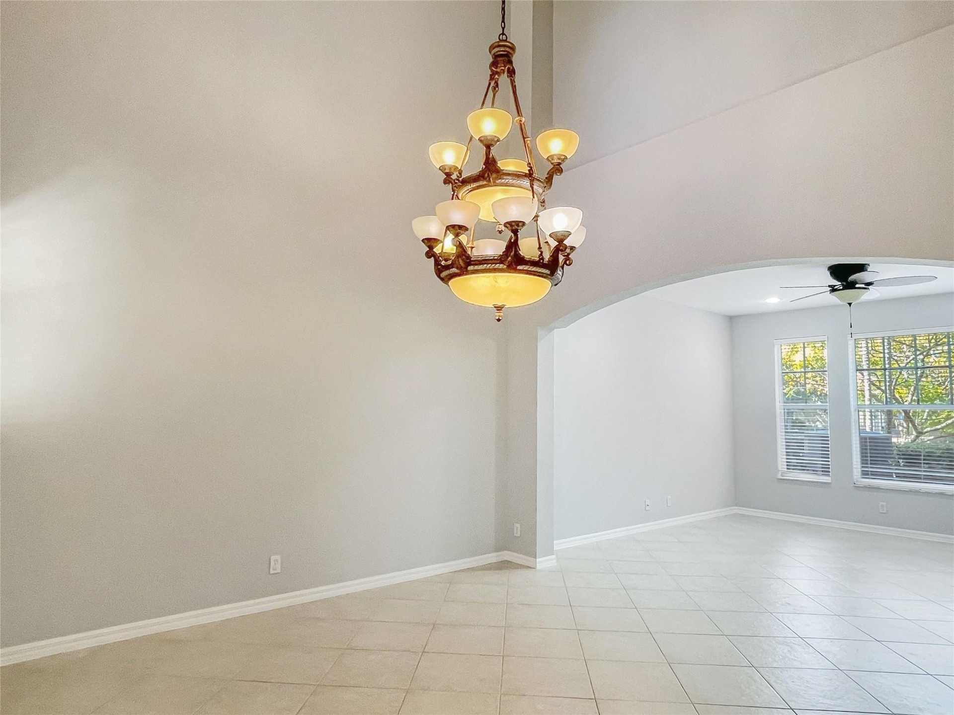 High-ceilinged room featuring a multi-tiered bronze-finish chandelier, arched architectural detailing, tile flooring, and a ceiling fan