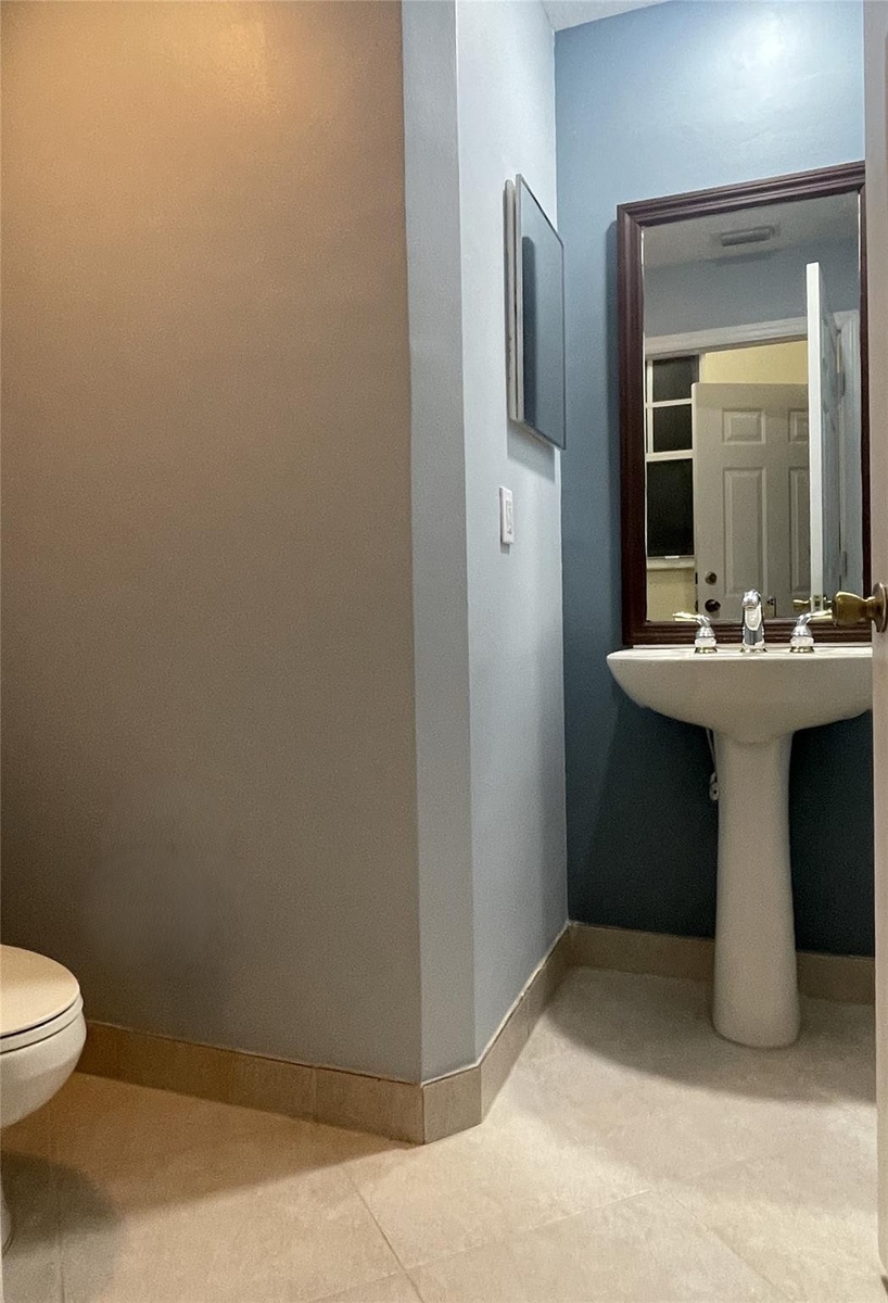 Pedestal sink featuring a polished chrome faucet, wall-mounted dark wood-frame mirror, and neutral-tone tile flooring