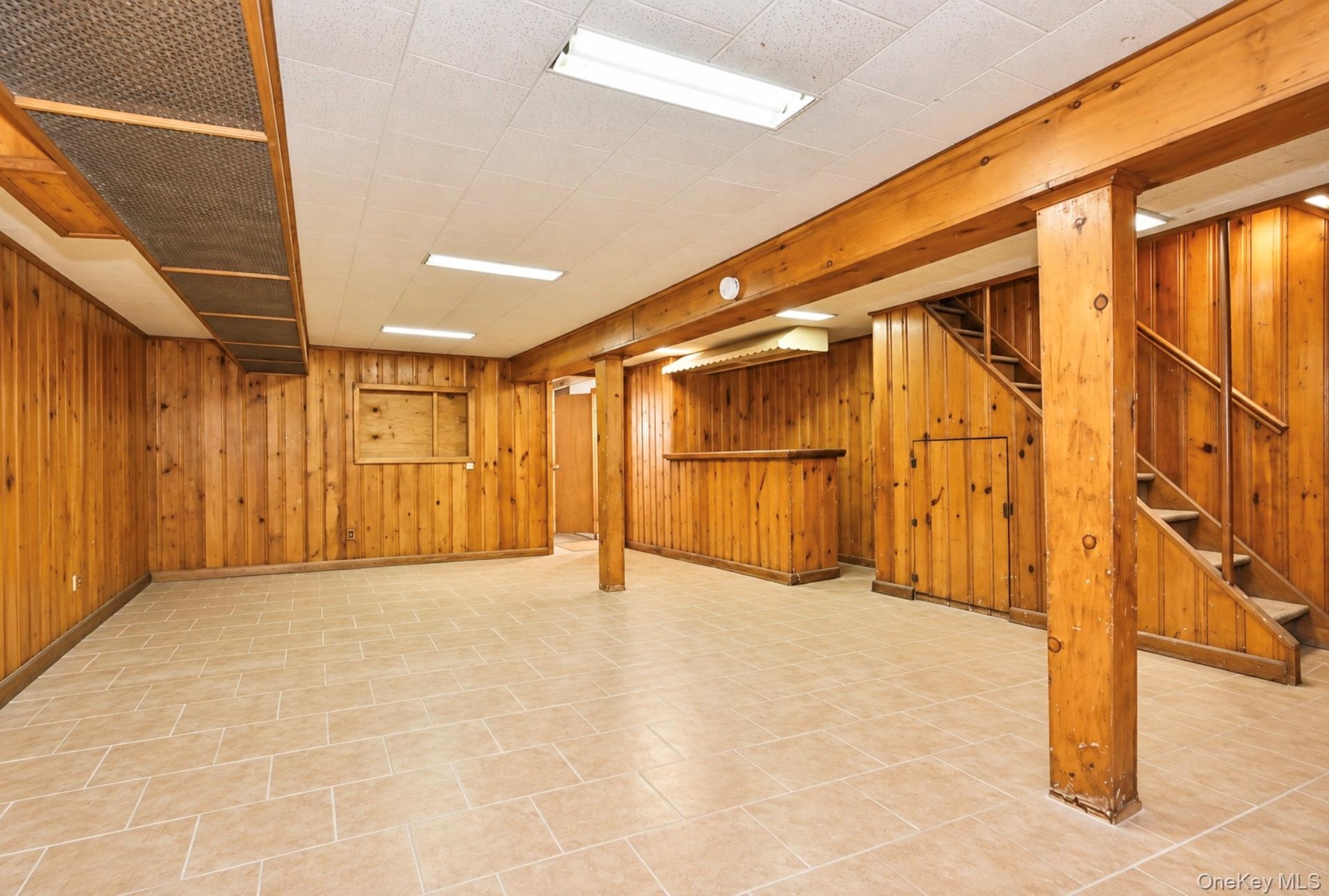 Interior space featuring wood-panelled walls, ceramic tile flooring, a built-in wooden bar, an open wooden staircase, and exposed structural support p