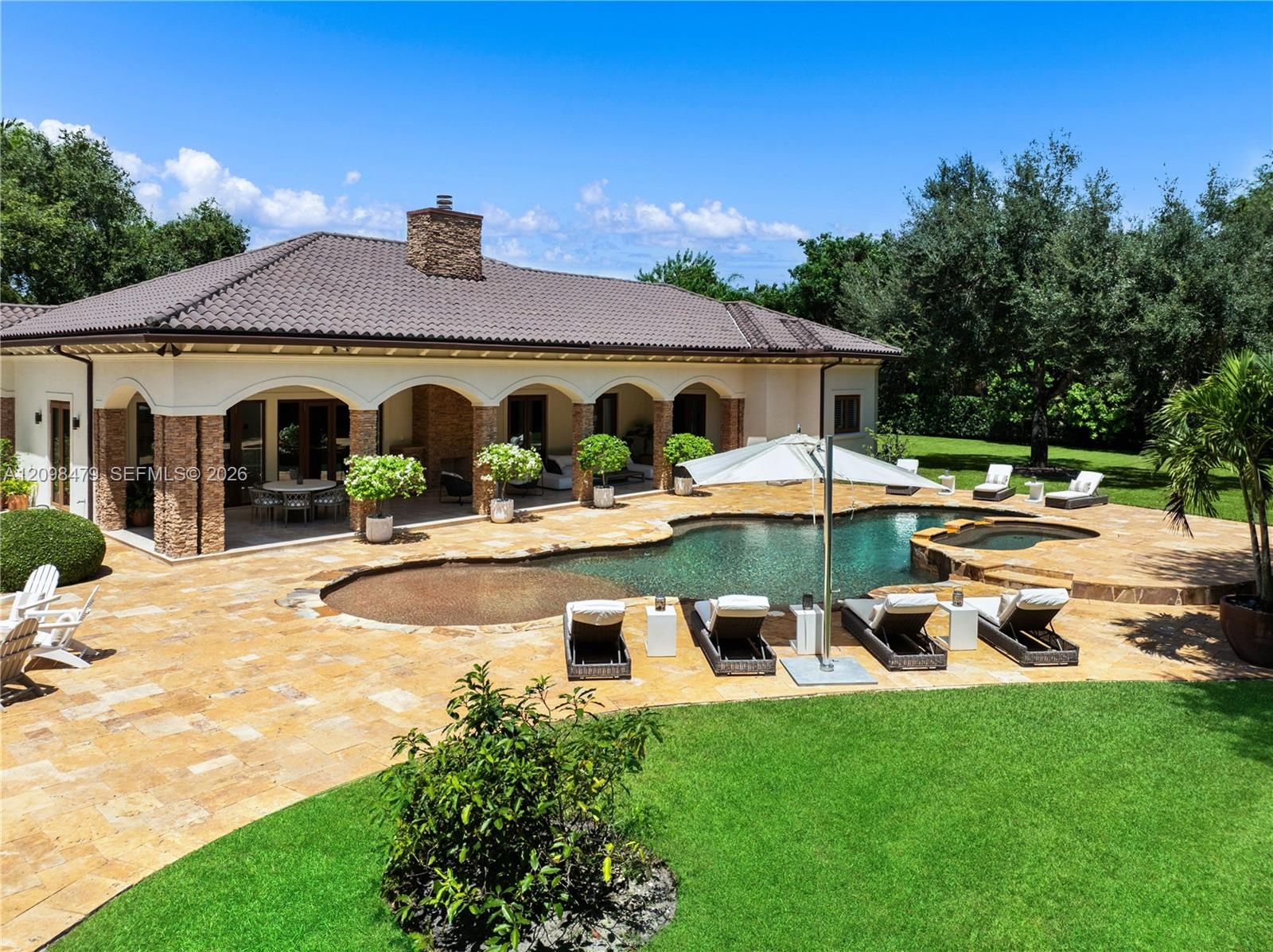 Expansive stone patio featuring a freeform swimming pool with integrated spa, a covered loggia with stone pillar accents, and a manicured lawn